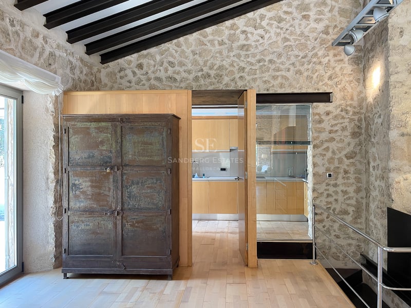 Intérieur avec murs en pierre naturelle, parquet en bois clair, poutres foncées et vue sur une cuisine moderne.