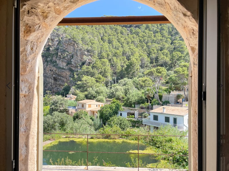 Vue à travers une arche en pierre sur une vallée verdoyante, des falaises rocheuses et des maisons traditionnelles.