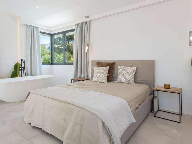 Chambre minimaliste avec baignoire blanche, literie neutre et fenêtres d'angle donnant sur la verdure.
