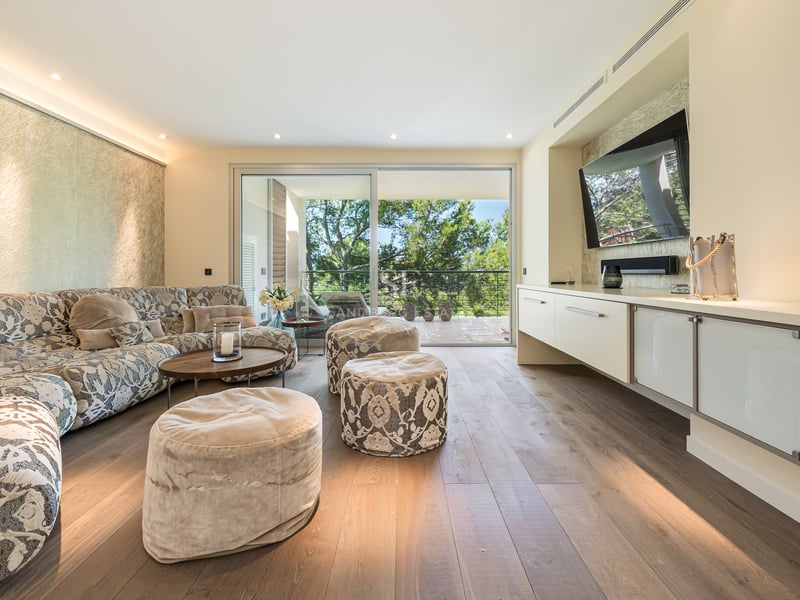 Vaste salon avec canapé à motifs, parquet en chêne et grandes baies vitrées ouvrant sur une terrasse boisée.