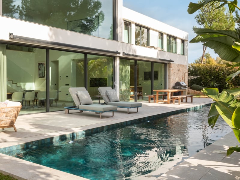 Piscine turquoise à côté d'une villa blanche moderne avec de grandes baies vitrées et des transats.