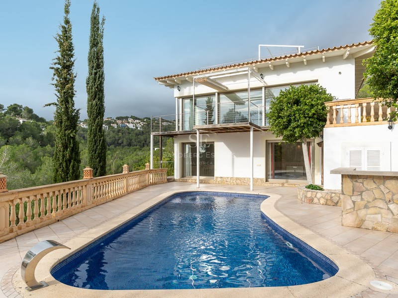 Une piscine de forme libre sur une terrasse en pierre à côté d'une villa blanche de deux étages avec vue sur la forêt.
