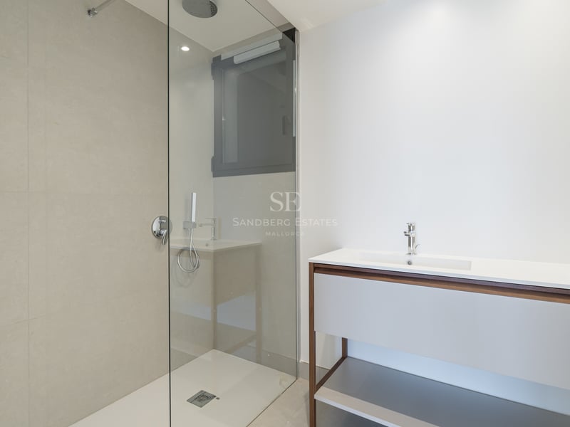 Baño moderno con ducha de cristal, azulejos beige y un mueble de lavabo elegante en blanco y madera.