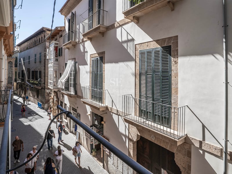 Vue depuis un balcon sur une rue étroite avec des bâtiments en pierre claire, des volets verts et des passants.