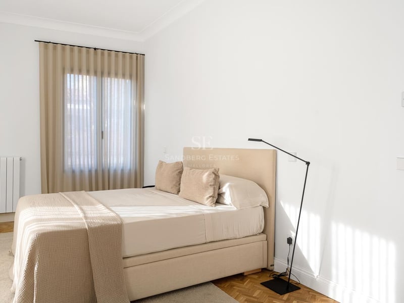 Chambre lumineuse avec un lit beige, des murs blancs et un parquet en bois chaleureux.