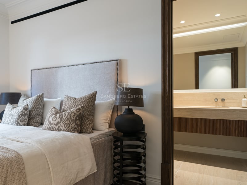 Modern bedroom featuring a tufted headboard and a view into a private bathroom with travertine stone vanity.