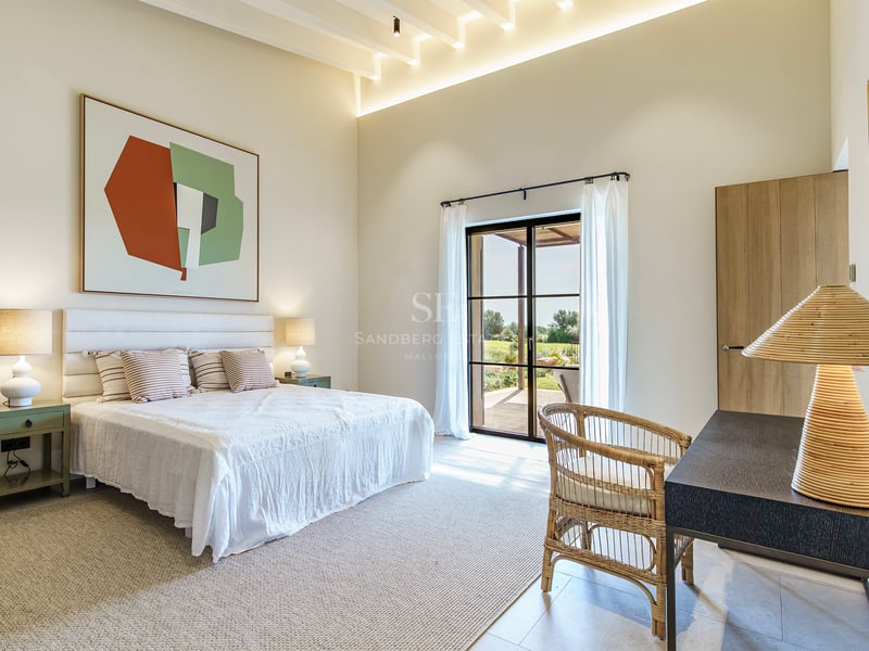 Dormitorio luminoso con techos altos, cama blanca, silla de mimbre y grandes puertas de cristal al jardín.