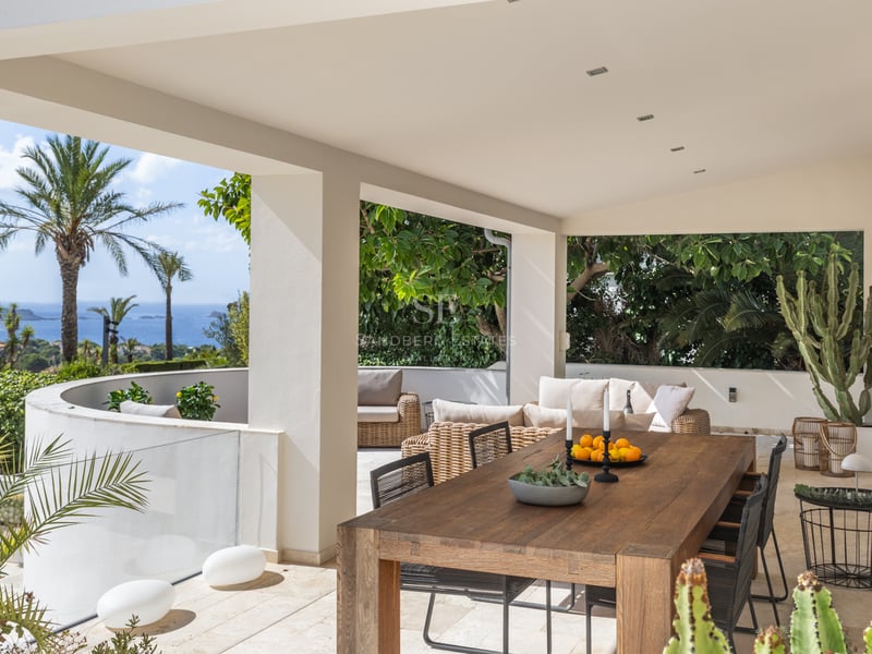 Terrasse couverte avec grande table en bois, canapés en rotin et vue sur la mer Méditerranée et les palmiers.