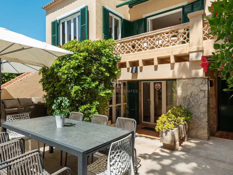 Terrasse en pierre avec salle à manger extérieure grise, parasol blanc et façade de villa aux volets verts.