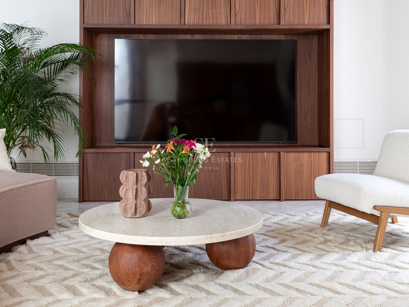 Salón contemporáneo con mueble de TV en nogal, mesa de centro redonda de travertino y sillón de bouclé blanco.