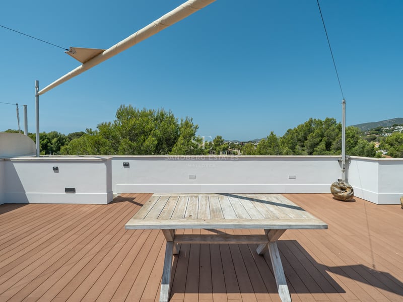 Grande terrasse de toit avec plancher en bois, table rustique, voile d'ombrage et vue sur les pins et collines.