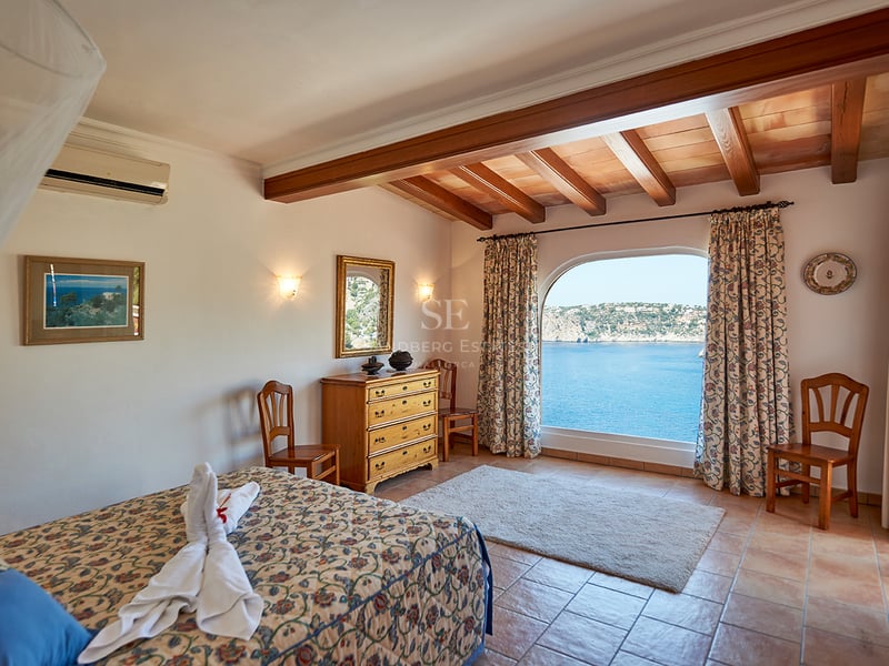Chambre avec sols en terre cuite, poutres apparentes et une grande fenêtre voûtée donnant sur la mer bleue.