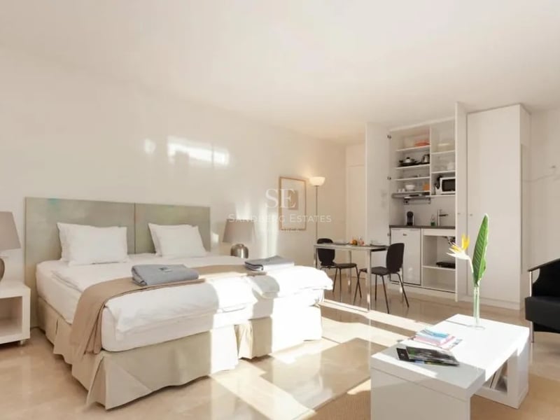 Studio lumineux avec lit blanc, sol en marbre et kitchenette dissimulée dans des placards blancs.