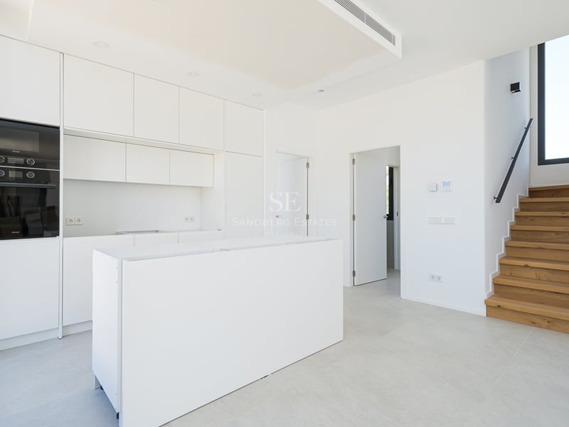 Cuisine moderne blanche avec îlot central, appareils intégrés et escalier en bois menant à l'étage.