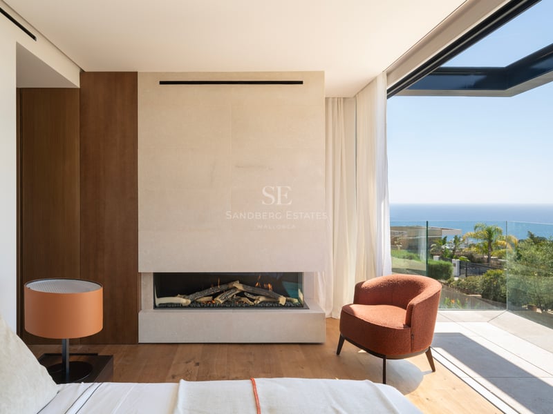 Chambre de maître moderne avec cheminée en pierre, fauteuil orange et baies vitrées ouvrant sur une terrasse vue mer.