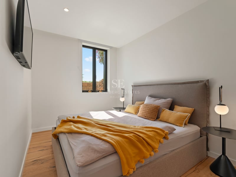Chambre moderne avec lit gris, jeté de lit moutarde, parquet en bois et fenêtre lumineuse.