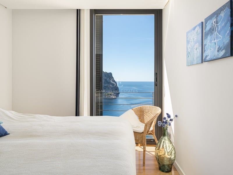 Chambre lumineuse avec literie blanche, oreiller bleu et baies vitrées ouvrant sur un paysage côtier méditerranéen.