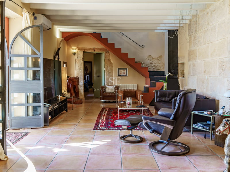 Salon lumineux avec murs en pierre apparente, sol en terre cuite, poutres en bois et fauteuils en cuir.