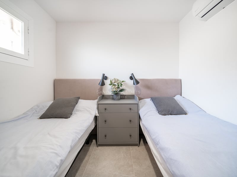 Chambre lumineuse avec deux lits simples, têtes de lit beiges, commode grise centrale et liseuses murales noires.