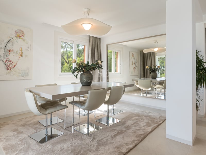 Salle à manger moderne avec table aux tons neutres, chaises chromées, grand miroir mural et vue sur le jardin.