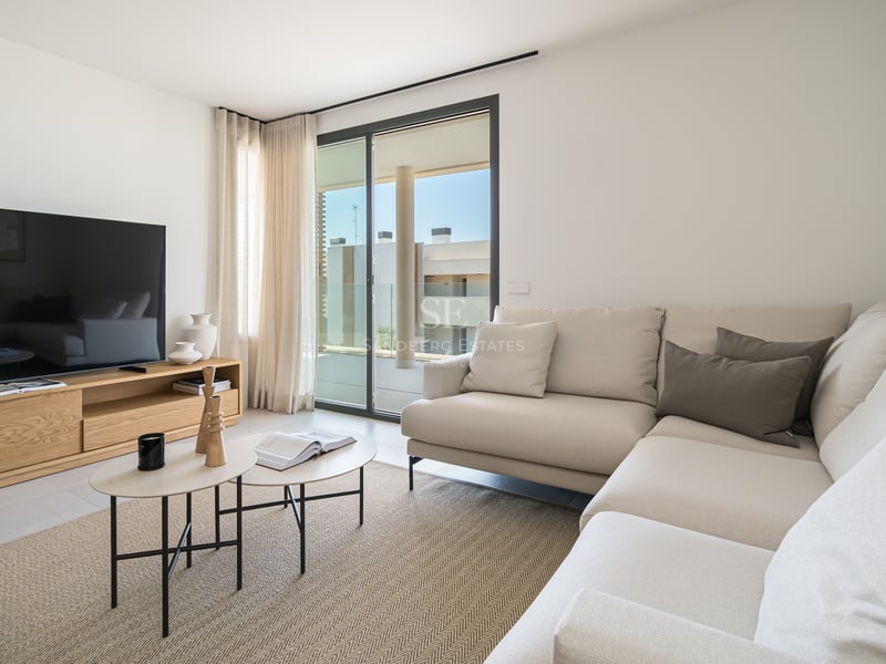 Salón luminoso con sofá en L color crema, mueble de TV en roble y puertas correderas hacia una terraza soleada.