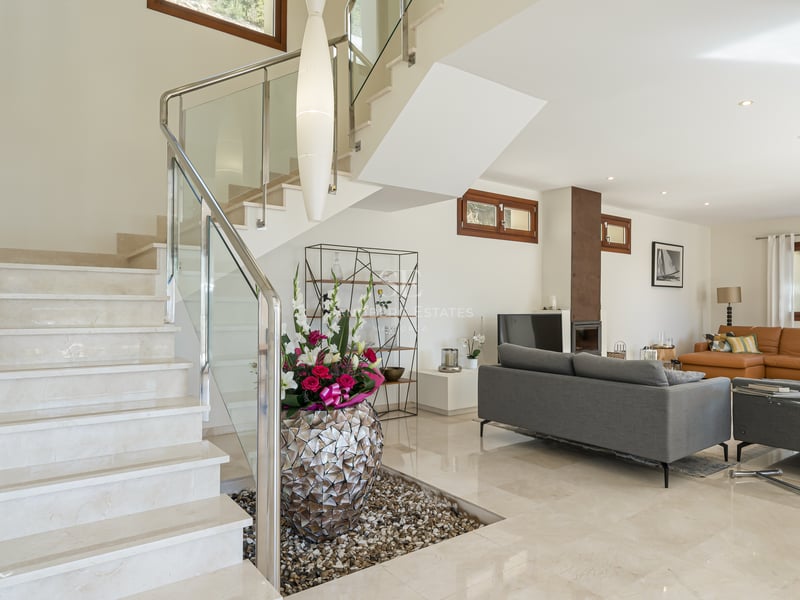 Salon lumineux à aire ouverte avec escalier en marbre crème, balustrades en verre et canapés gris.