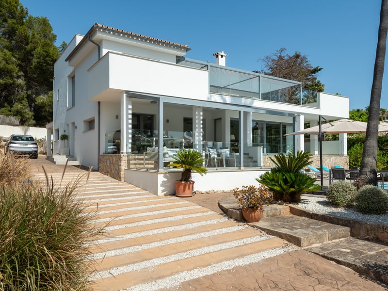 Villa blanche moderne de deux étages avec terrasses vitrées, accents en pierre et jardin soigné sous un ciel bleu clair.