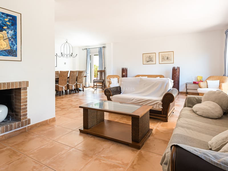 Grand salon avec sol en terre cuite, meubles en osier, cheminée en brique et coin repas adjacent.