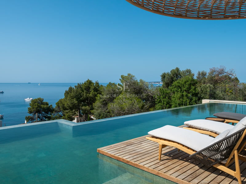Piscine à débordement avec terrasse en bois et bains de soleil face à la mer bleue et aux pins.