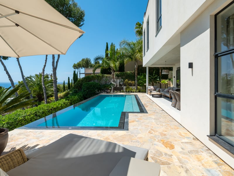 Une piscine turquoise sur un patio en pierre à côté d'une villa blanche moderne, surplombant la mer.
