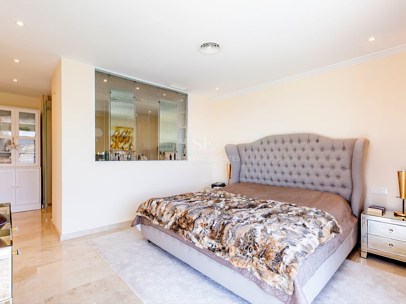 Chambre spacieuse avec tête de lit capitonnée grise, sol en marbre, tables de chevet en miroir et rangements.