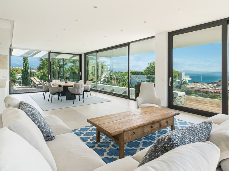 Salon spacieux avec canapés blancs, table basse en bois et grandes baies vitrées s'ouvrant sur une terrasse vue mer.
