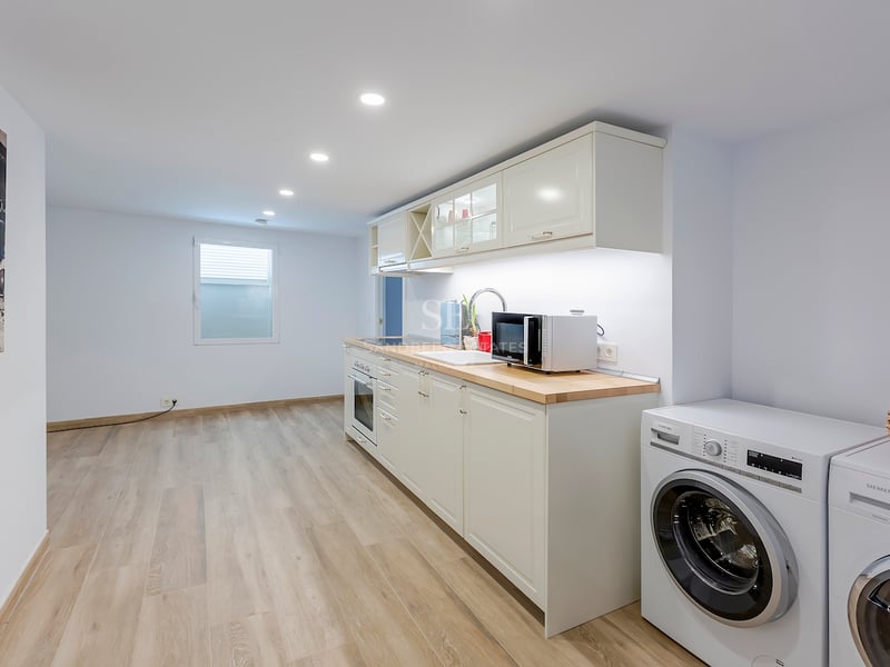 Cuisine moderne lumineuse avec placards blancs, plans de travail en bois clair et lave-linge et sèche-linge intégrés.