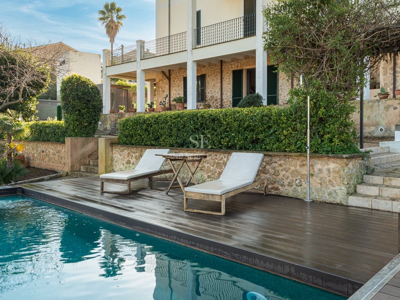 Piscine turquoise avec terrasse en bois sombre, chaises longues et façade en pierre rustique.