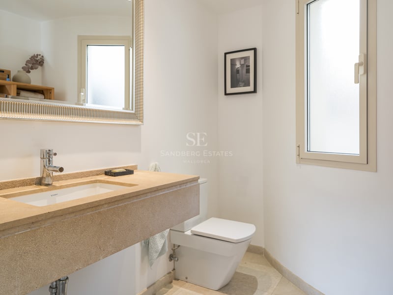 Salle de bain lumineuse avec vasque en pierre beige, grand miroir au cadre argenté, toilettes blanches et fenêtre dépolie.