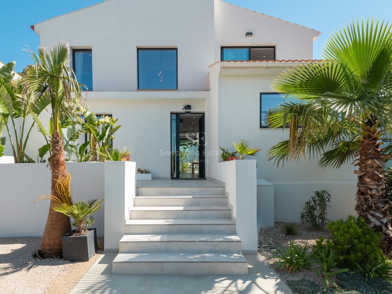 Extérieur d'une villa blanche avec un escalier en pierre claire, une porte vitrée et une végétation tropicale.