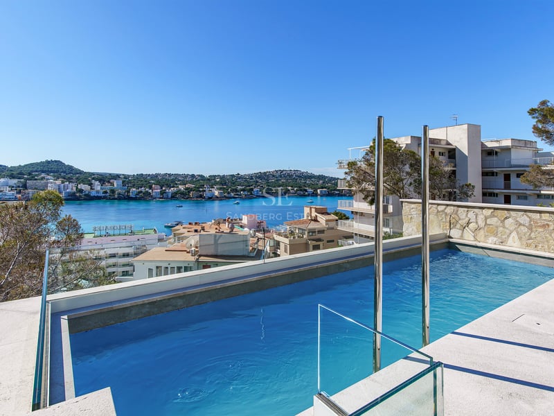Piscine moderne avec garde-corps en verre surplombant une baie méditerranéenne et une ville côtière sous un ciel bleu.