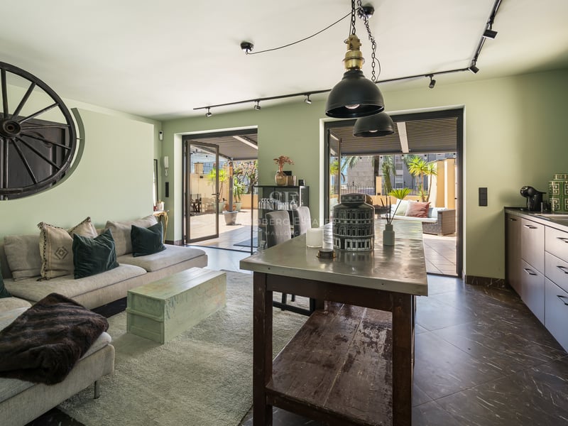 Intérieur d'un salon vert sauge et cuisine design s'ouvrant sur une terrasse privée ensoleillée.