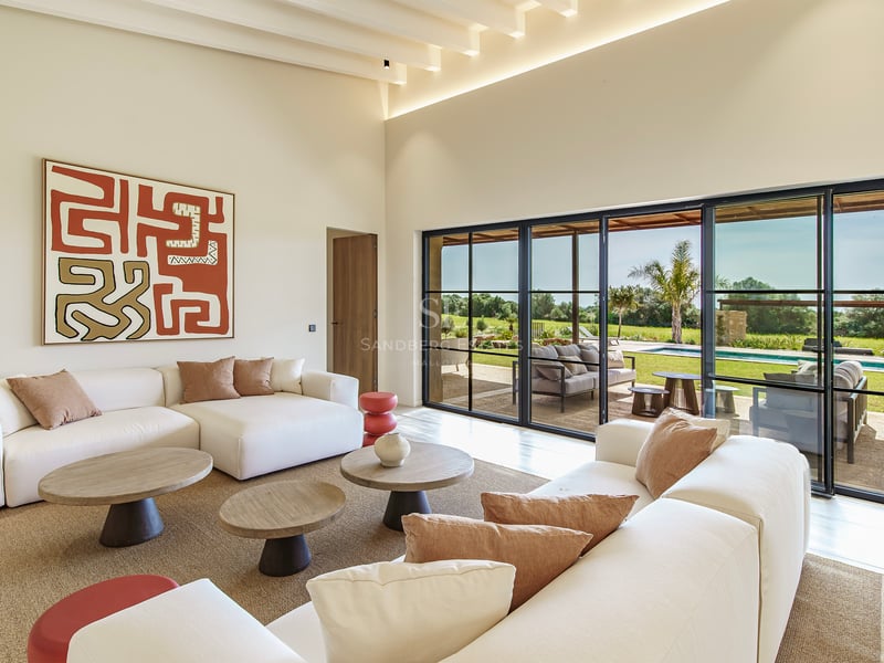 Salón luminoso con sofás blancos, mesas de madera y grandes ventanales con vistas al jardín y la piscina.
