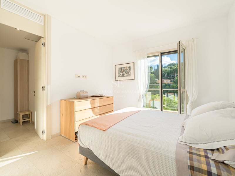 Chambre lumineuse avec lit, commode en bois clair et porte de balcon ouverte donnant sur la verdure.