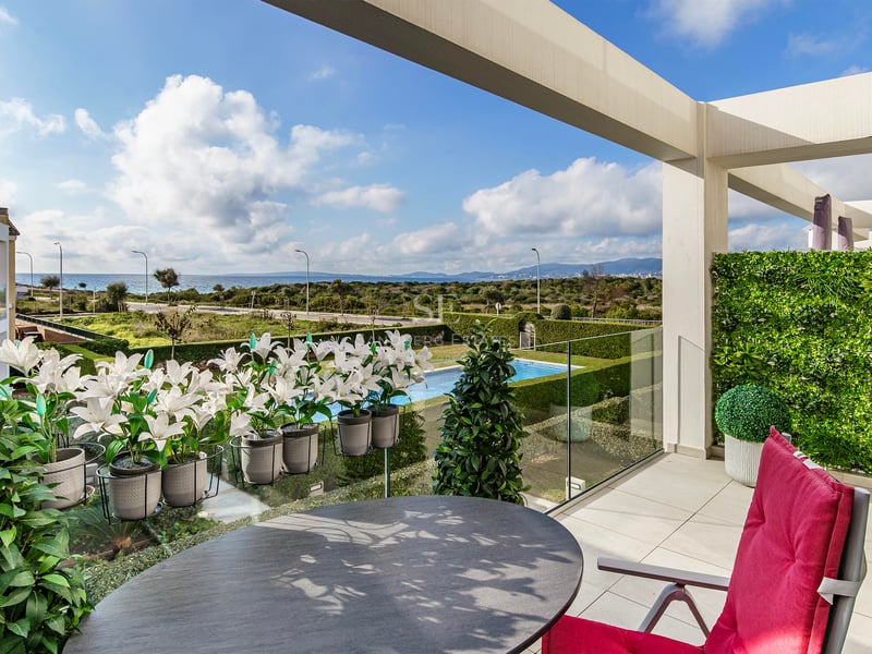 Terrasse de luxe avec table ronde, chaise rouge et garde-corps en verre surplombant jardin, piscine et mer.