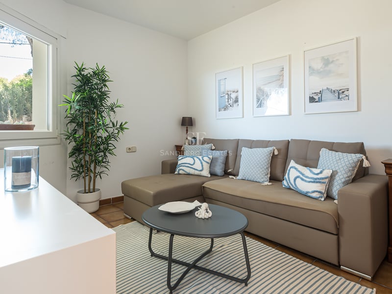 Un salon lumineux avec un canapé en L taupe, des coussins bleus, un tapis à rayures et des tableaux sur le thème de la mer.
