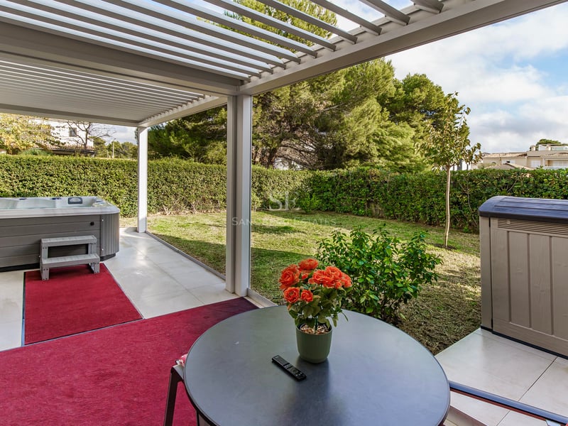 Terrasse avec pergola blanche, jacuzzi gris, tapis rouges et vue sur un jardin verdoyant avec haies.