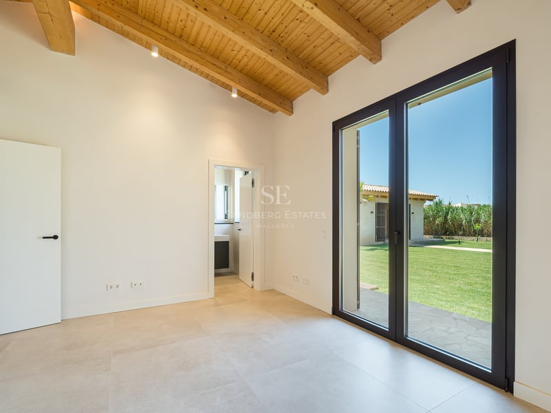 Dormitorio moderno vacío con techos altos de vigas de madera y grandes puertas de cristal hacia un jardín verde.