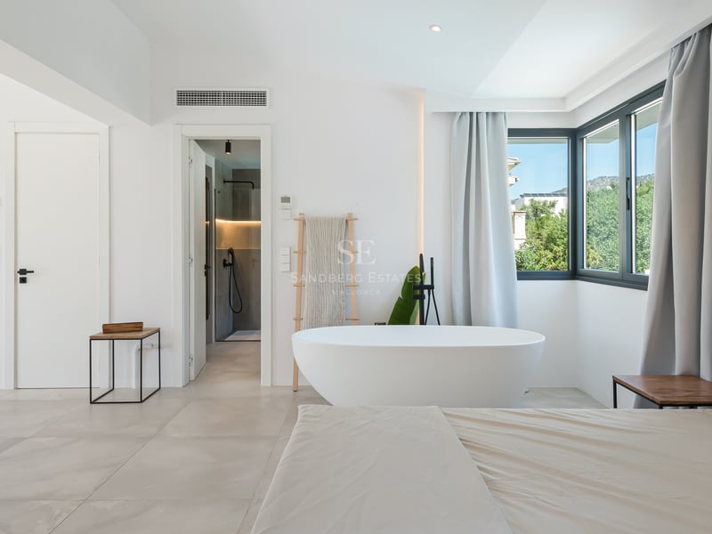 Grande chambre blanche avec baignoire îlot ovale, rideaux gris et salle de bain attenante visible à l'arrière-plan.