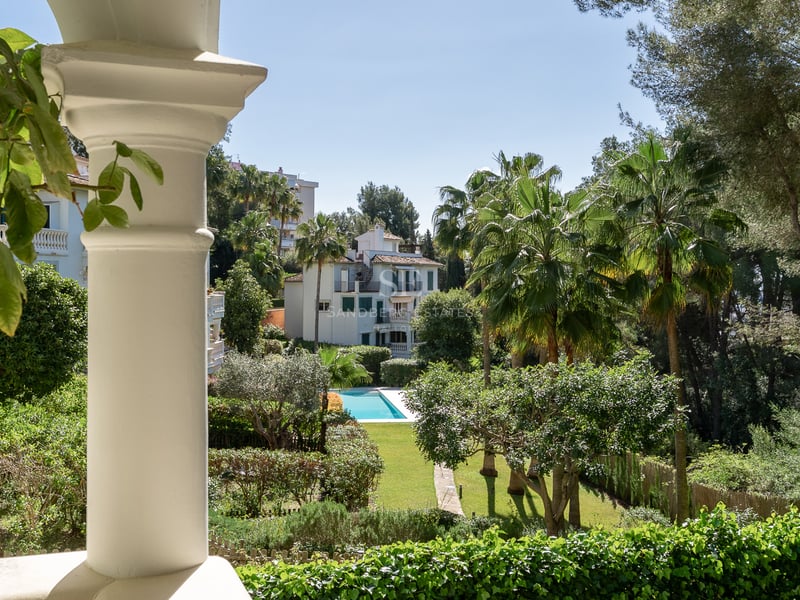 Vue depuis une colonne blanche sur un jardin verdoyant avec une piscine bleue et des palmiers sous un ciel clair.