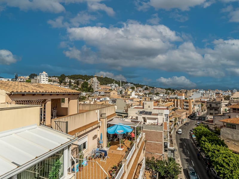 Vue surélevée des toits d'une ville méditerranéenne, une rue bordée d'arbres et des montagnes au loin.