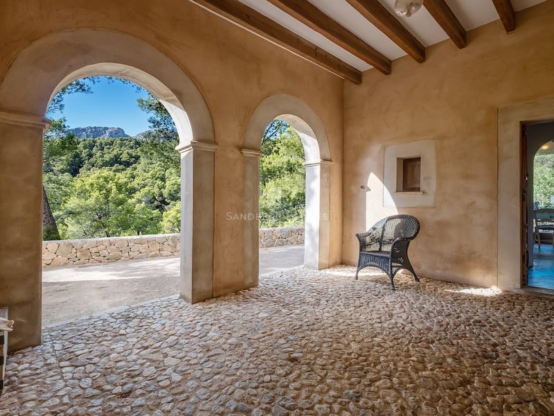Terrasse rustique avec arches, poutres en bois, sol en galets et vue sur la forêt.