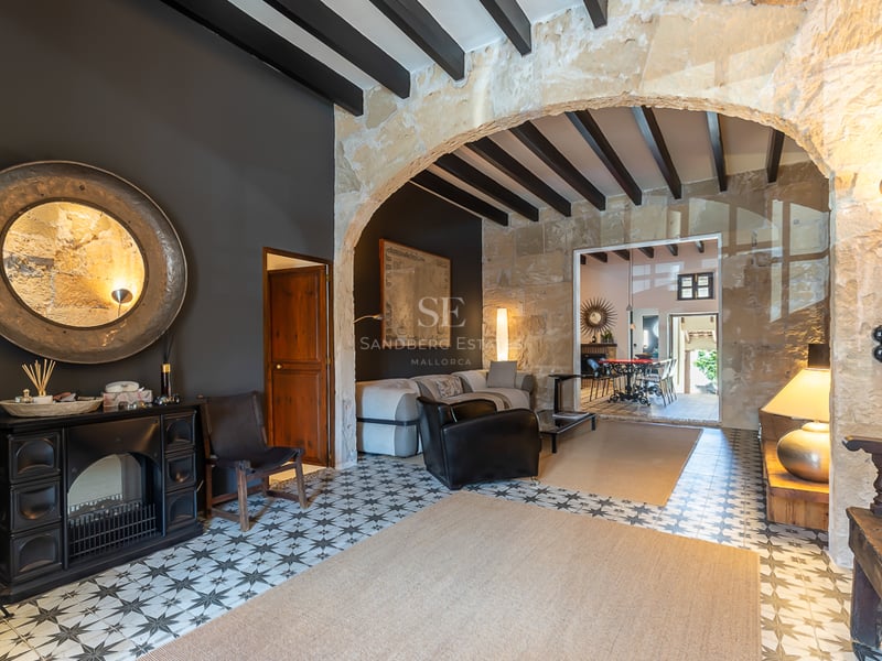 Interior view of a living room featuring a large natural stone archway, dark ceiling beams, and patterned hydraulic floor tiles.