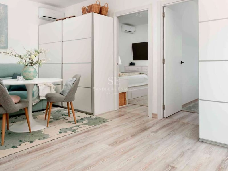 Espace de vie lumineux avec table ronde blanche, chaises grises, parquet clair et vue sur la chambre attenante.